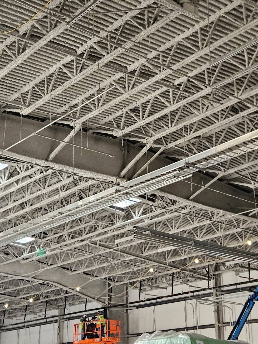 Spray-applied fire-resistive material installed on open-web bar joists and the metal deck above, with the crew working from a lift