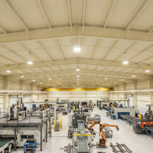 Interior of a large industrial manufacturing facility with spray foam insulation coating the arched metal roof deck and structural steel framing above active production equipment.