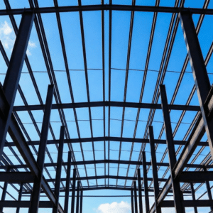 Bare structural steel frame of a commercial building under construction, showing wide-flange columns and curved roof purlins silhouetted against a blue sky — unprotected steel prior to spray-applied fireproofing application