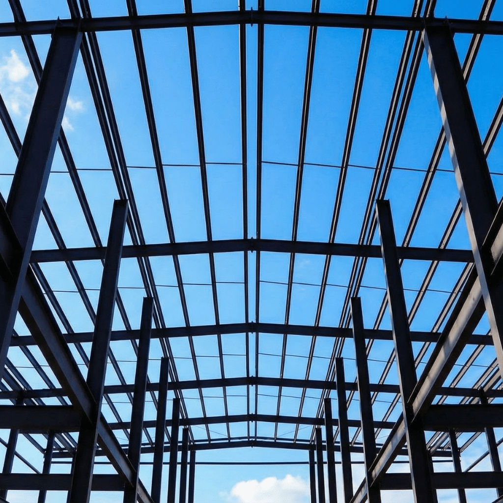 Fire Ratings for Structural Steel: 1-Hour to 4-Hour Explained 1 Bare structural steel frame of a commercial building under construction, showing wide-flange columns and curved roof purlins silhouetted against a blue sky — unprotected steel prior to spray-applied fireproofing application