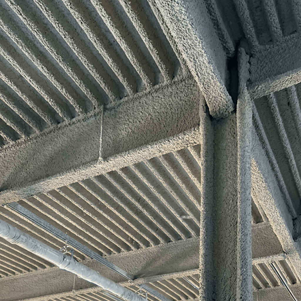 How Spray-Applied Fireproofing Is Applied: A Practical Process Guide for GCs 1 Close-up view of a commercial building ceiling with structural steel beams and corrugated metal deck fully coated in thick gray, concrete-like spray-applied fireproofing, with small pipes and conduit running below.