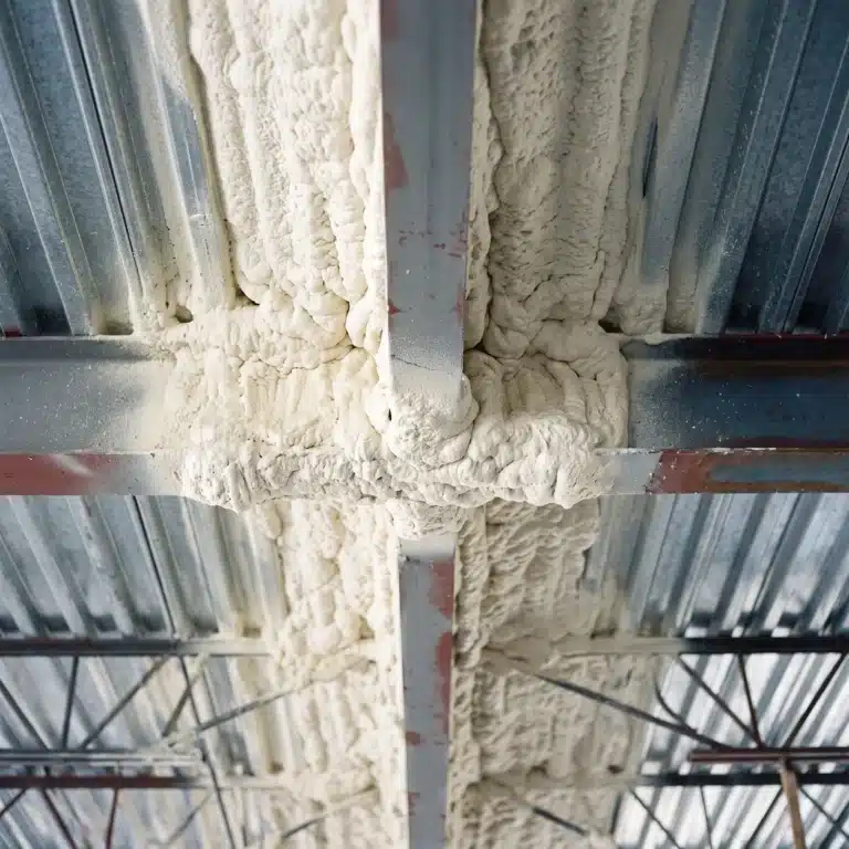 Agricultural insulation: off-white closed-cell spray foam coating the underside of a corrugated metal roof deck and packed around steel purlins